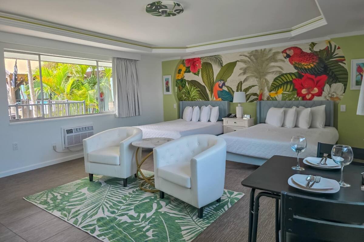 A modern hotel room with a large bed, two white armchairs, a small table, and a dining area.