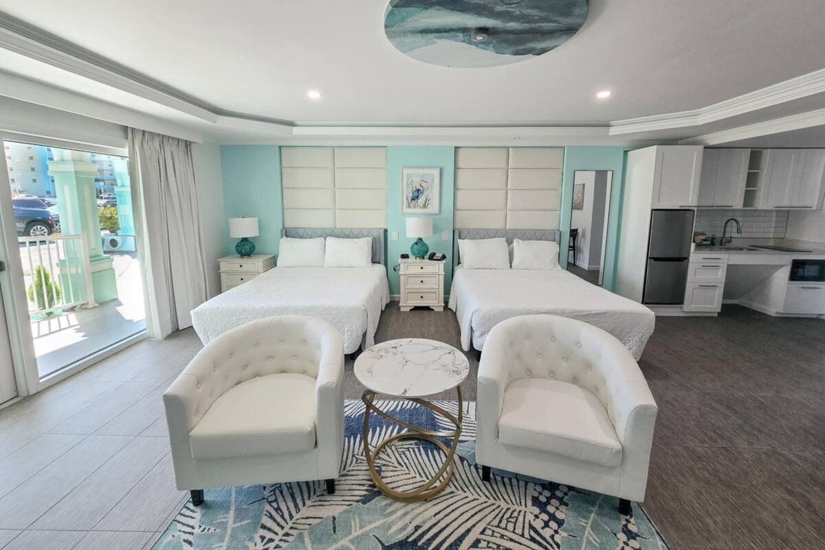 A hotel room with two beds, a small kitchen, a dining area, and a balcony with curtains.