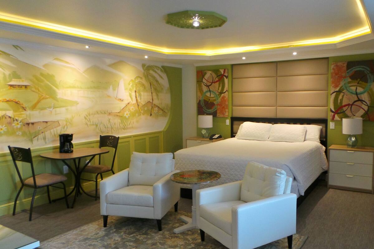 A modern hotel room with a large bed, two white armchairs, a small table, and a painting on the wall.