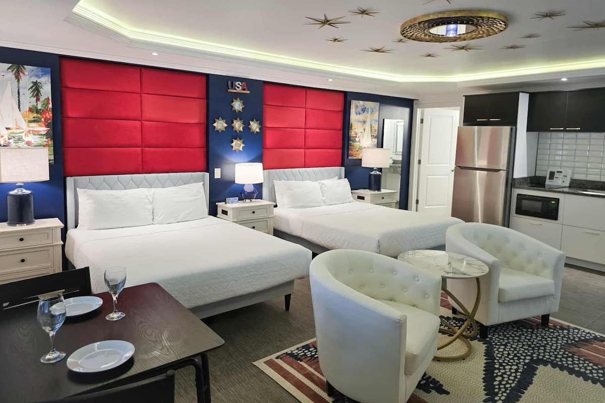 A hotel room with two beds, a dining table with plates and glasses, a sofa, a kitchen area with a refrigerator and microwave, and a ceiling with star decorations.