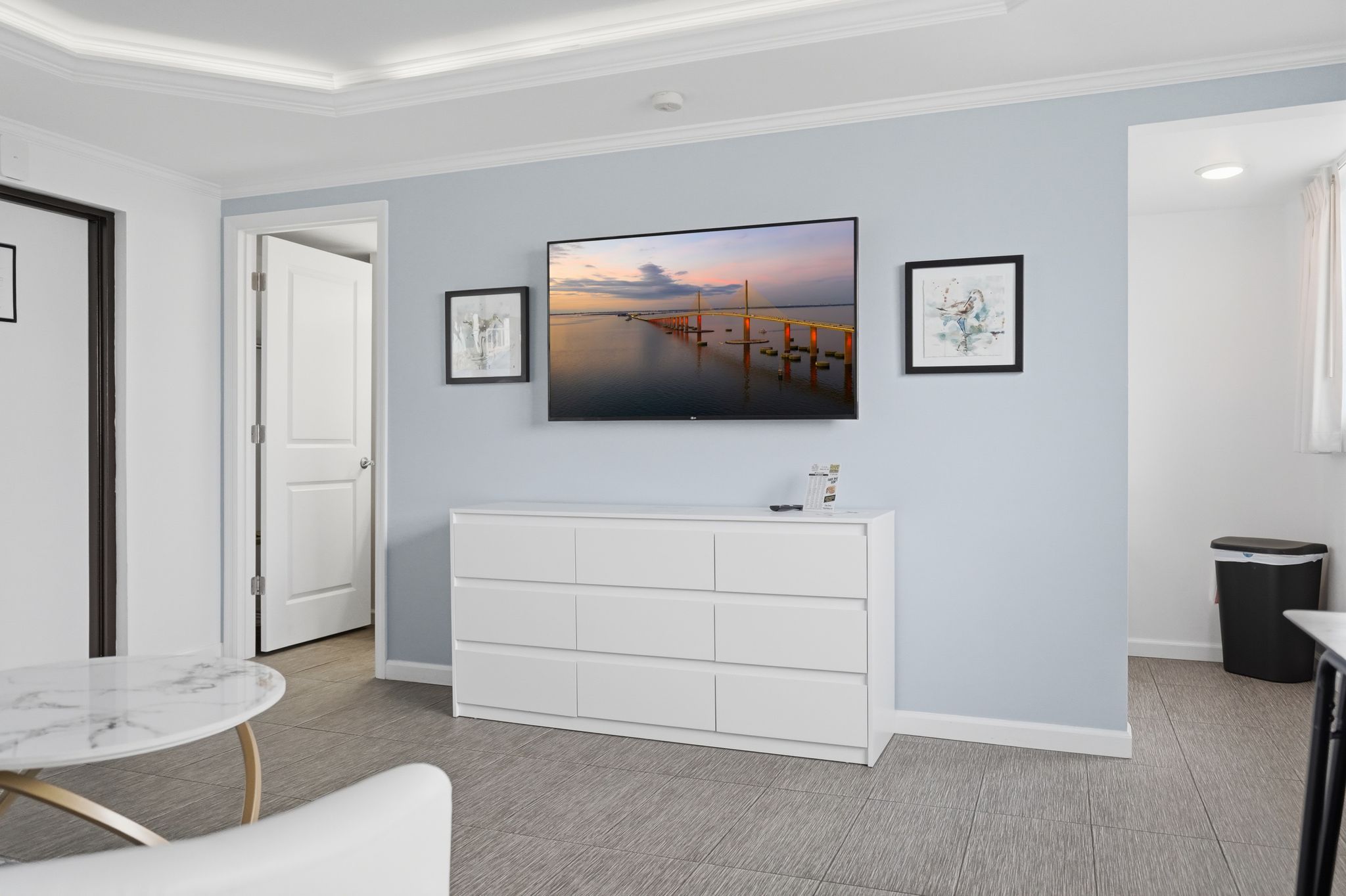 Una habitación moderna con un televisor de pantalla plana grande montado en la pared, una consola blanca, una mesa con cubierta de mármol y una puerta que conduce a otra habitación.