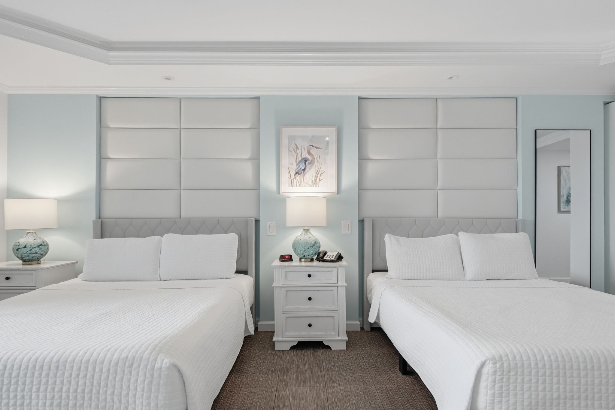 Quarto de hotel com duas camas, uma mesinha de cabeceira com abajur e um quadro na parede.