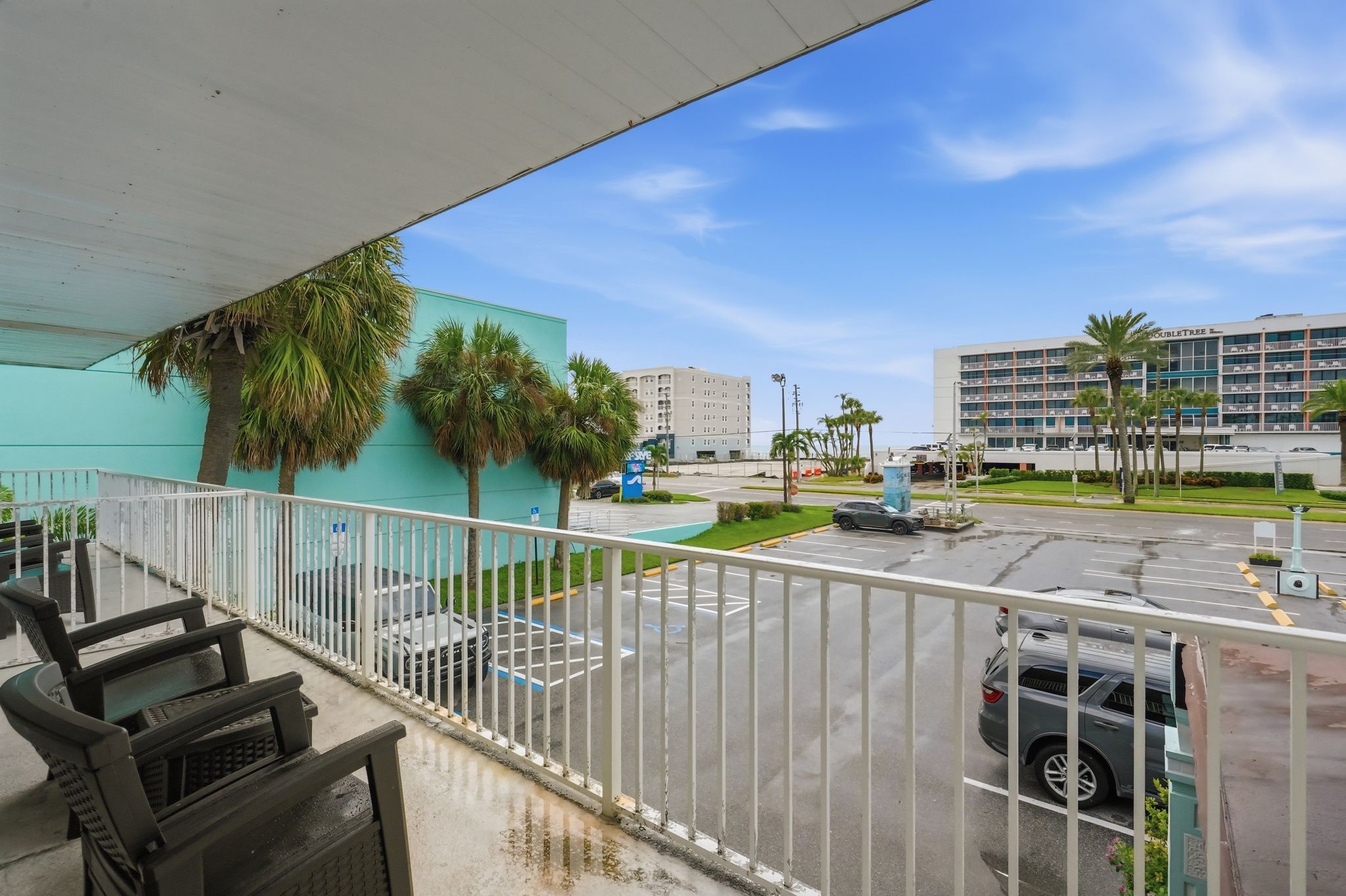 A balcony with a view of a parking lot, a building with a sign, and a palm tree.