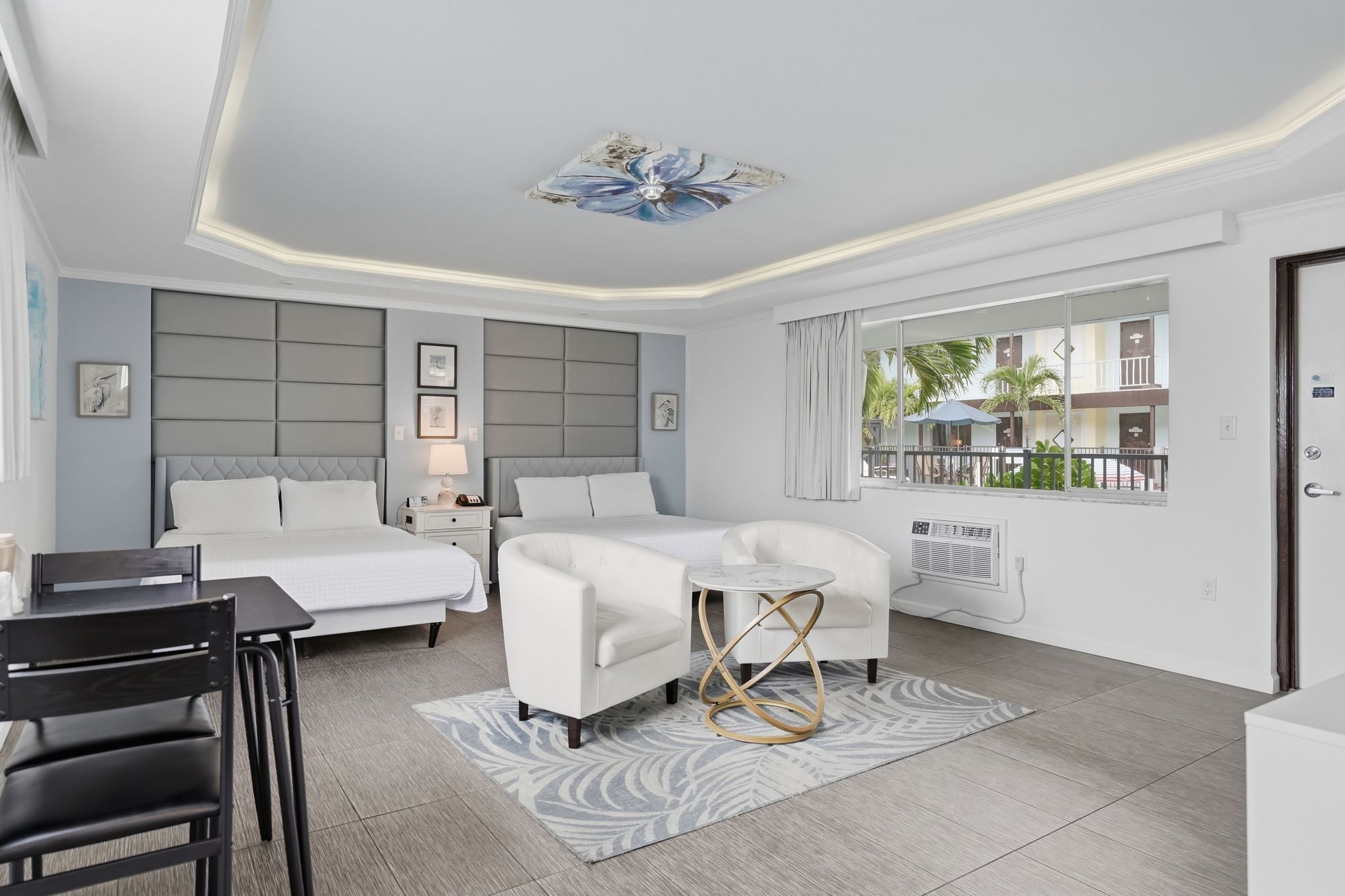 Habitación de hotel con una cama, dos sofás blancos, una mesita, una silla y una ventana con vistas a un edificio y árboles.