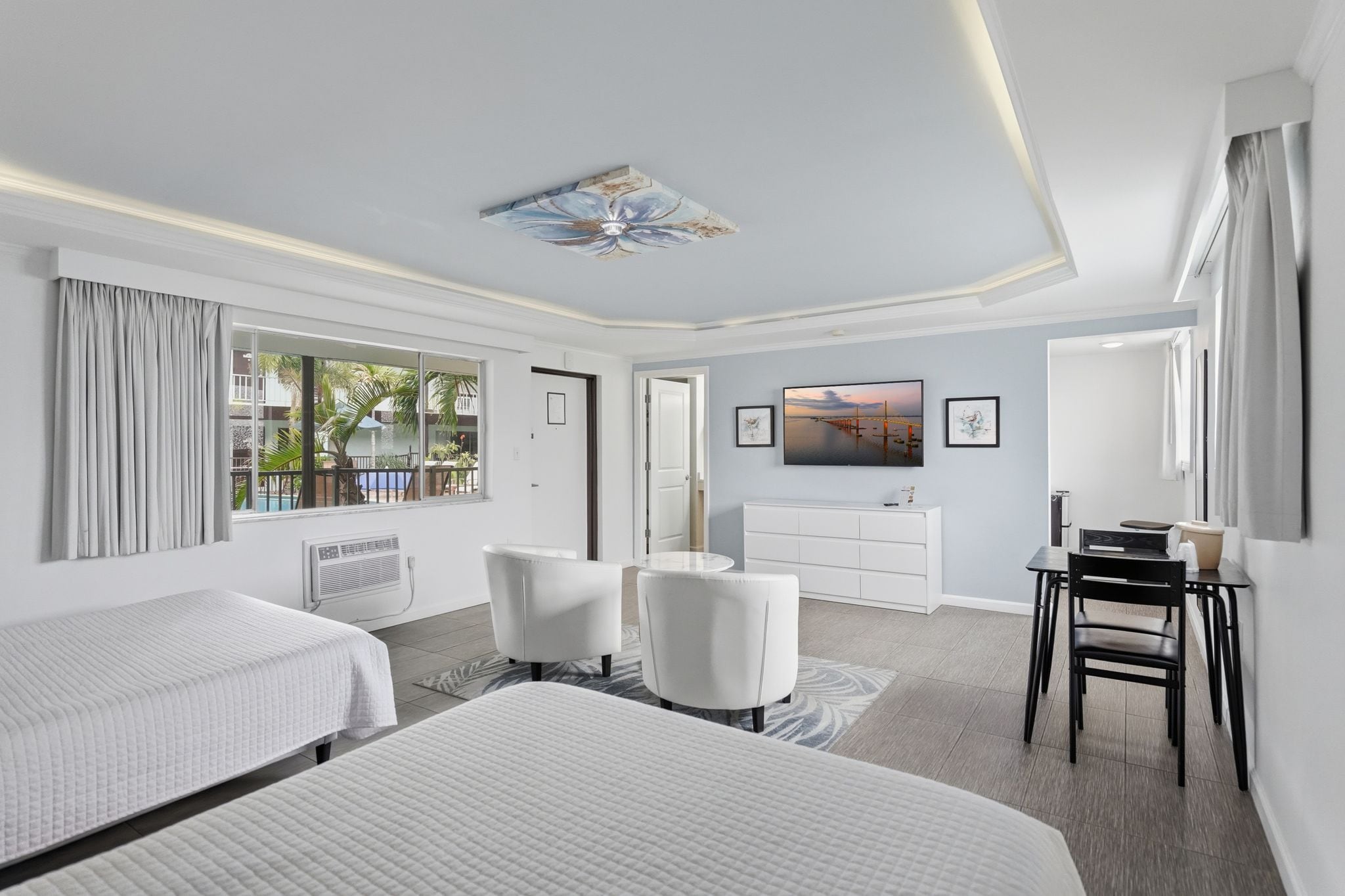 Habitación de hotel con dos camas, un escritorio, una silla, un televisor y vistas a una piscina y edificios.