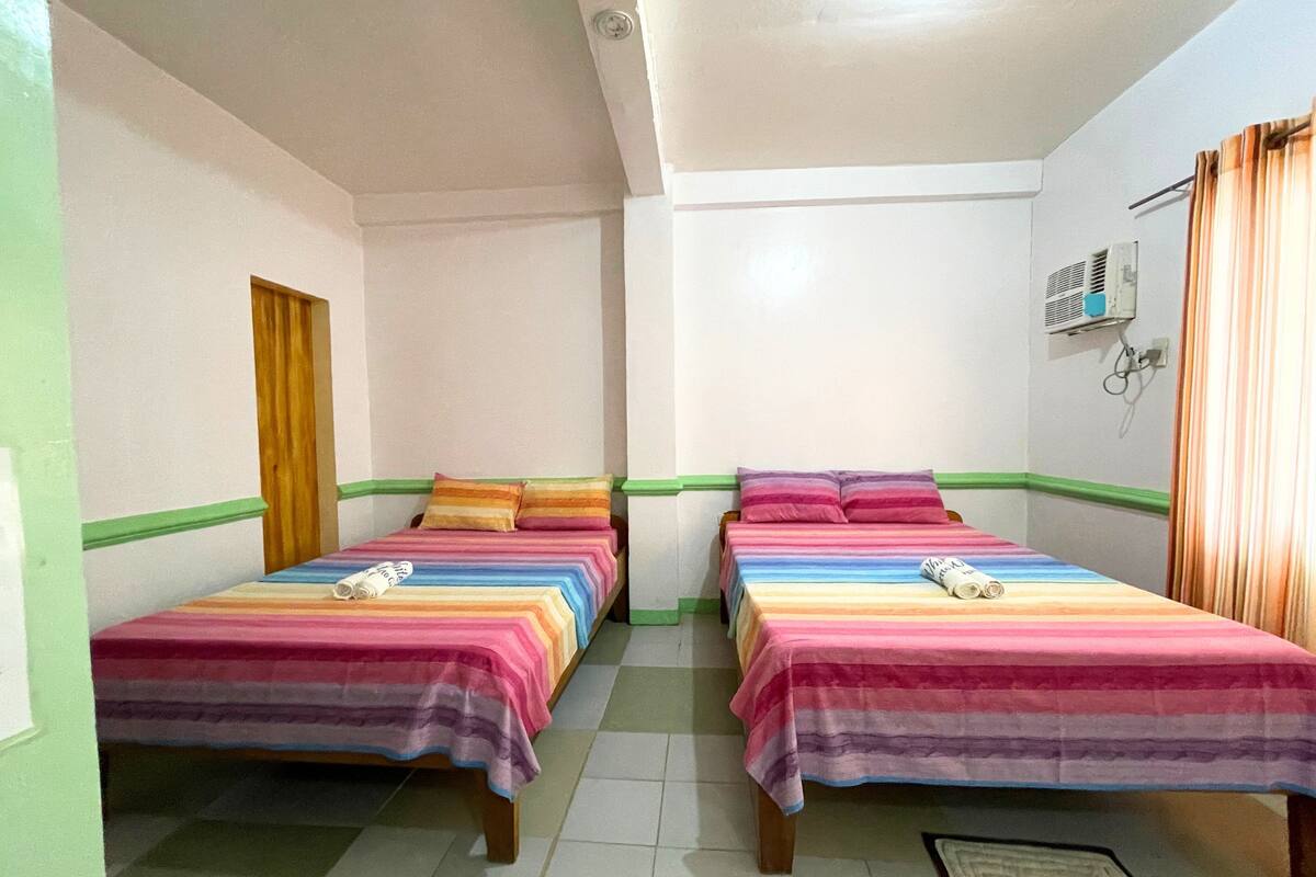 A room with two beds, each with colorful bedding. There is a window with curtains, a wall-mounted air conditioning unit, and a door.
