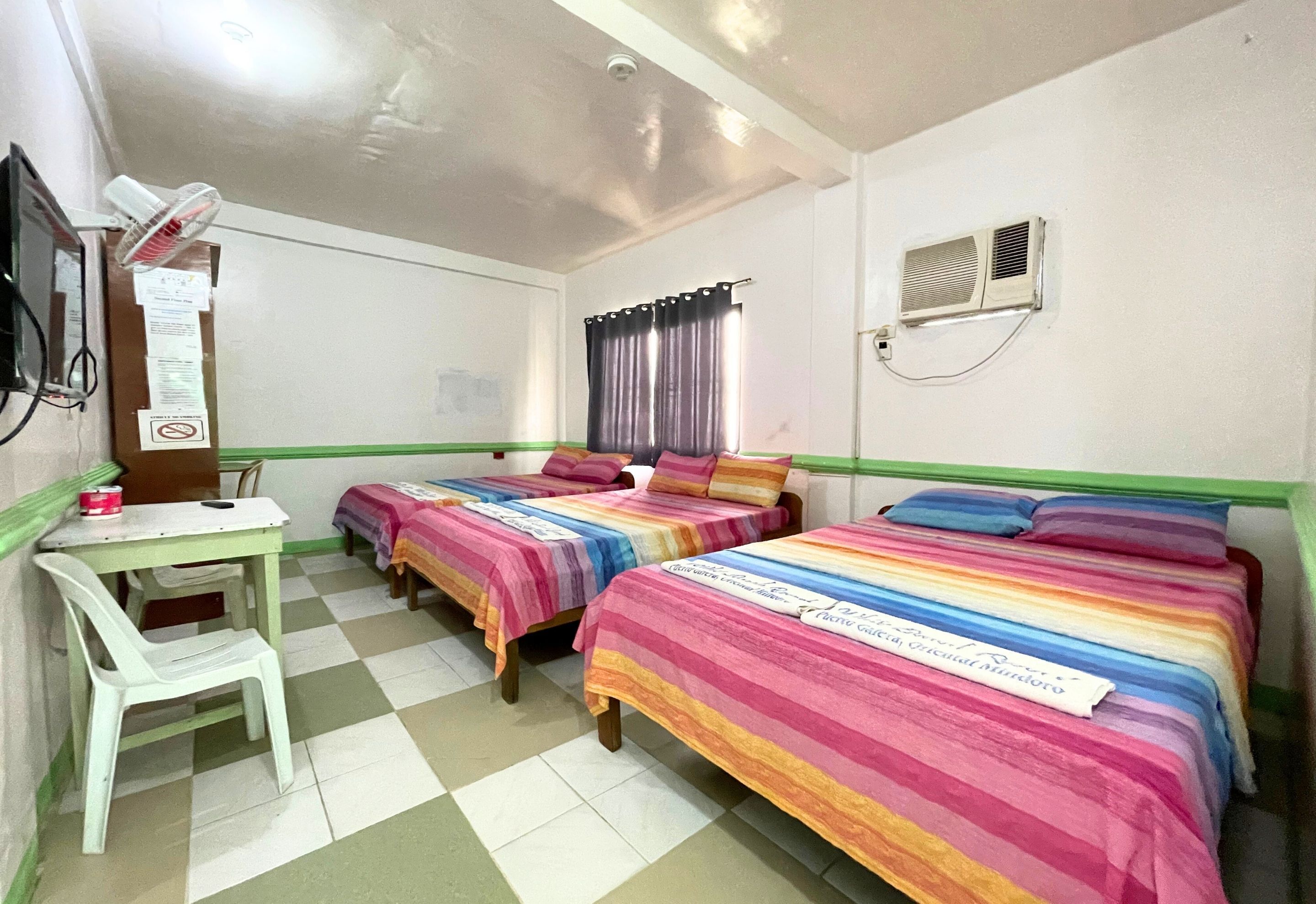 A room with two beds, a desk, a chair, a fan, a window with curtains, and an air conditioning unit.