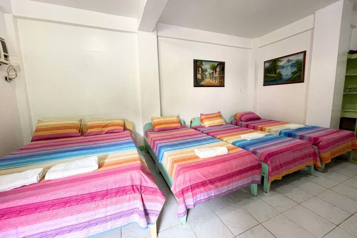A room with four beds, each with colorful striped bedding. There are two paintings on the walls.