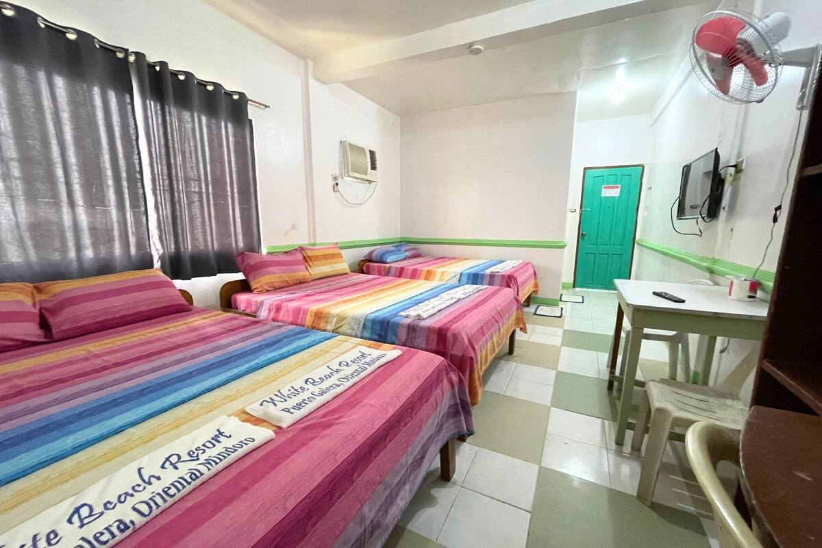 A hotel room with three beds, each with a striped bedspread, a desk with a chair, a green door, and a wall-mounted fan.