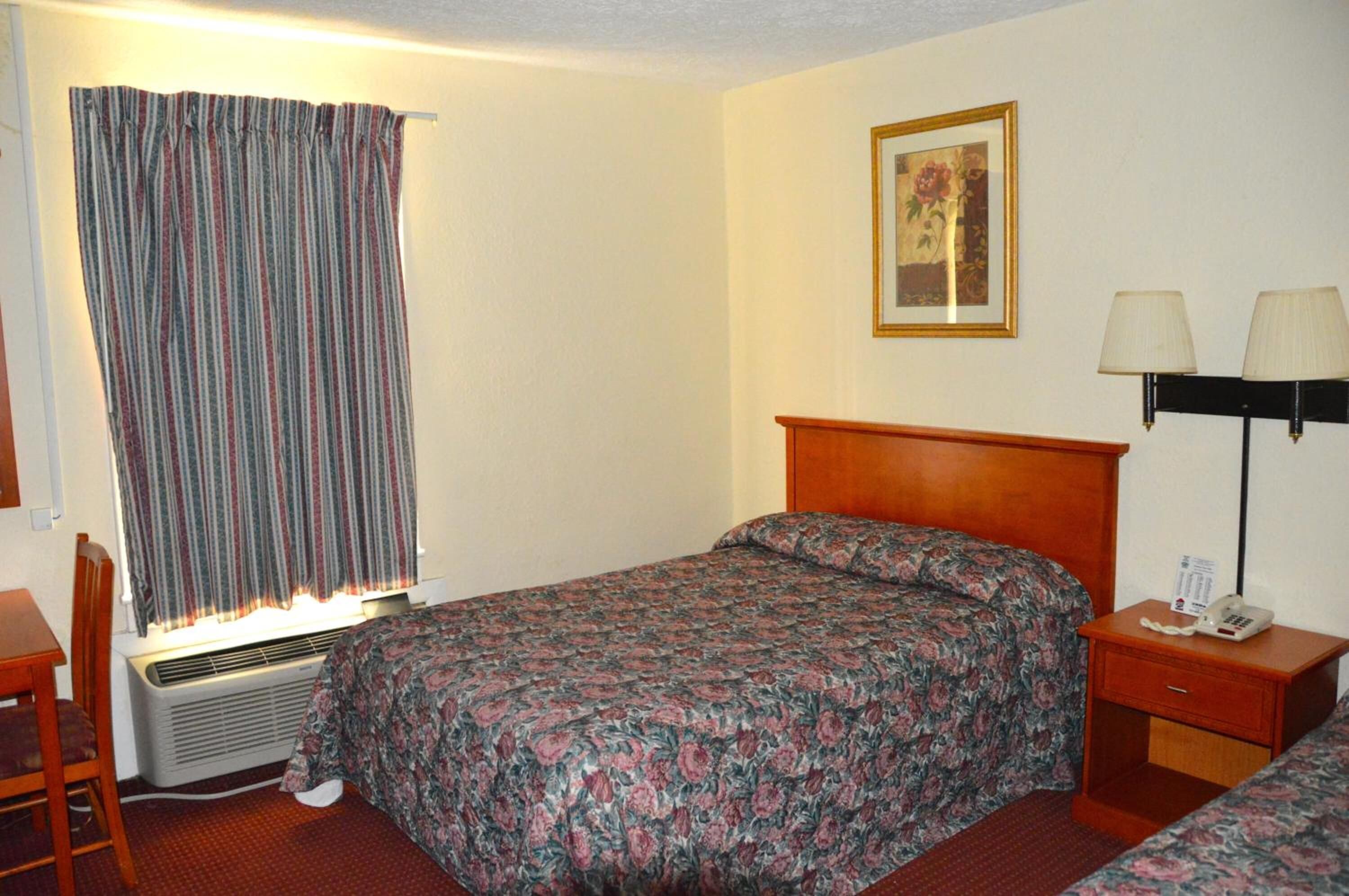 Ein Hotelzimmer mit Bett, Schreibtisch, Stuhl, Nachttisch mit Lampe und einem Fenster mit Vorhängen.