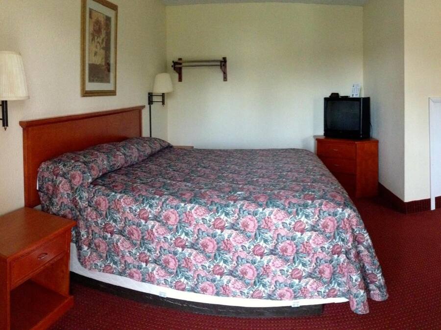Et hotelværelse med en enkelt seng, et natbord, et fjernsyn på et stativ og en lampe monteret på væggen.