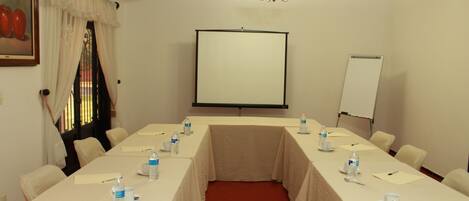 Un salón de conferencias con una larga mesa, sillas, botellas de agua y una pantalla de proyector.