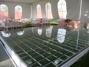 Un espace événementiel aménagé sous une tente, doté d’un plancher en verre, de chaises blanches et de tables disposées pour un événement.