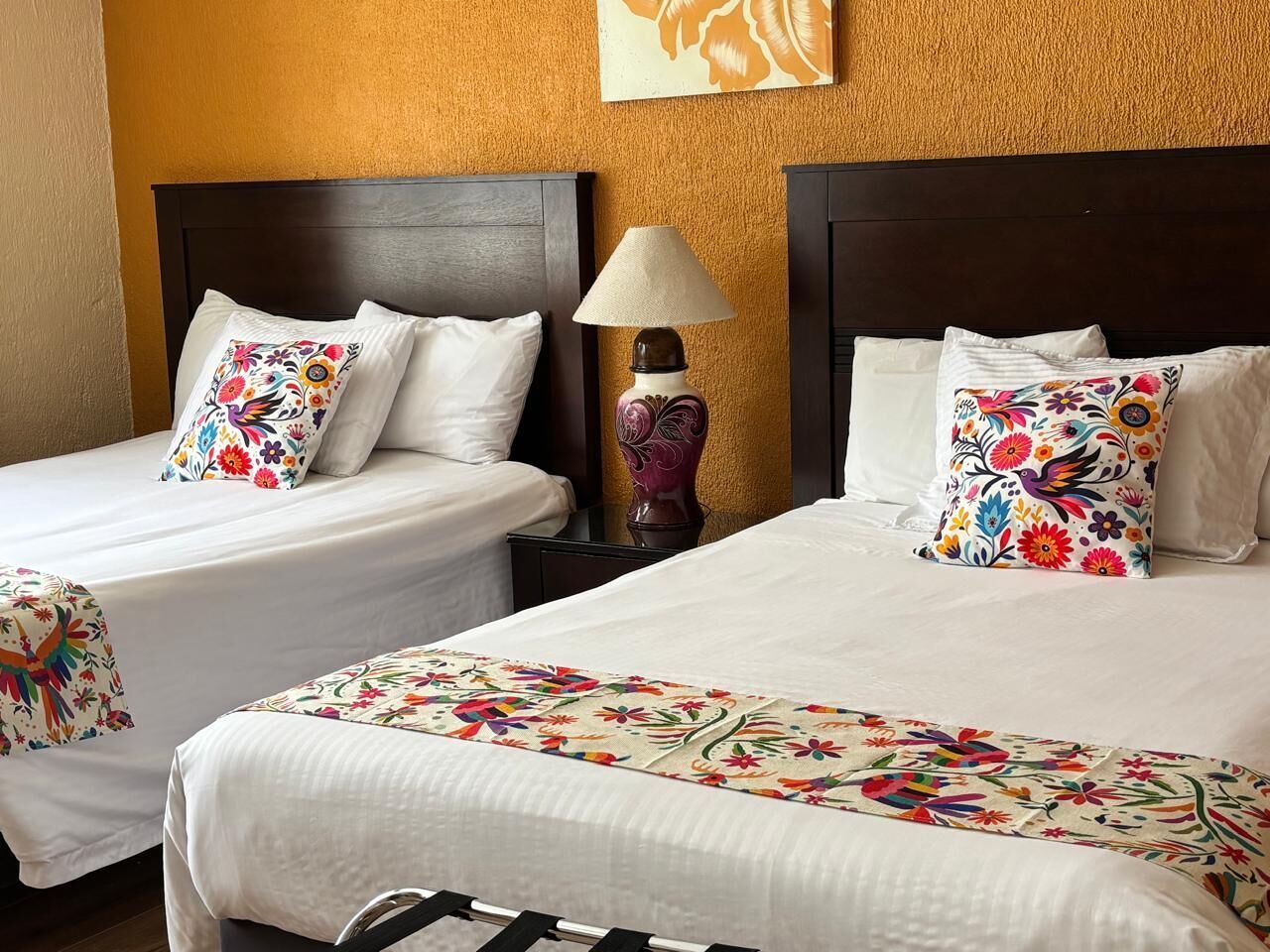 Ein Hotelzimmer mit zwei Betten, jedes mit blumigem Bettzeug und Kissen. Dazwischen steht ein Nachttisch mit einer Lampe.