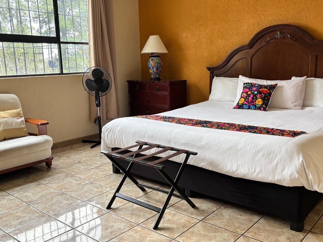A hotel room with a bed, a bedside table with a lamp, a chair, a fan, and a window with curtains.