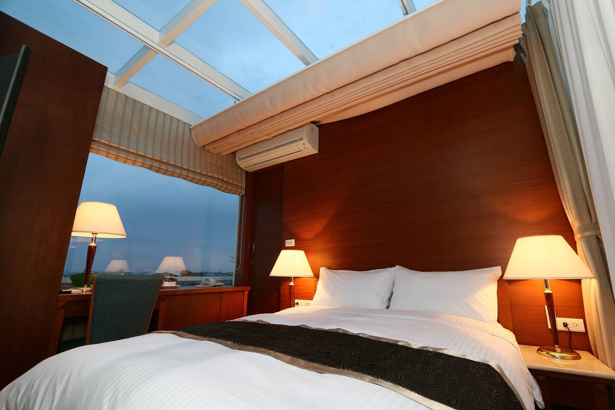 A hotel room with a large bed, two bedside lamps, a window with a view, and an air conditioning unit.