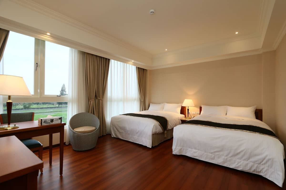 A hotel room with two beds, a desk with a lamp, a chair, and a view of the outdoors.