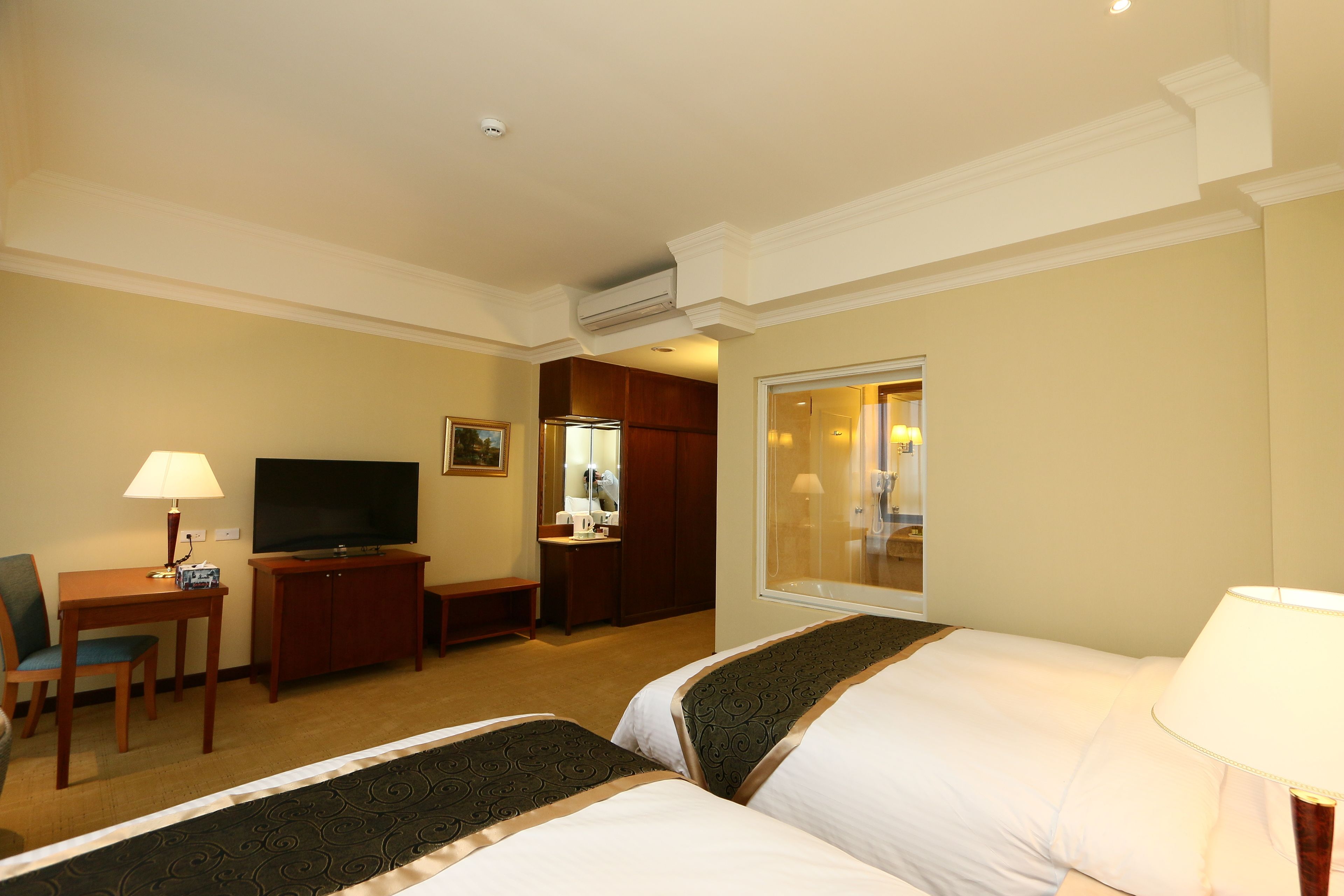 A hotel room with two beds, a television, a desk with a lamp, a chair, and a bathroom visible through a glass door.