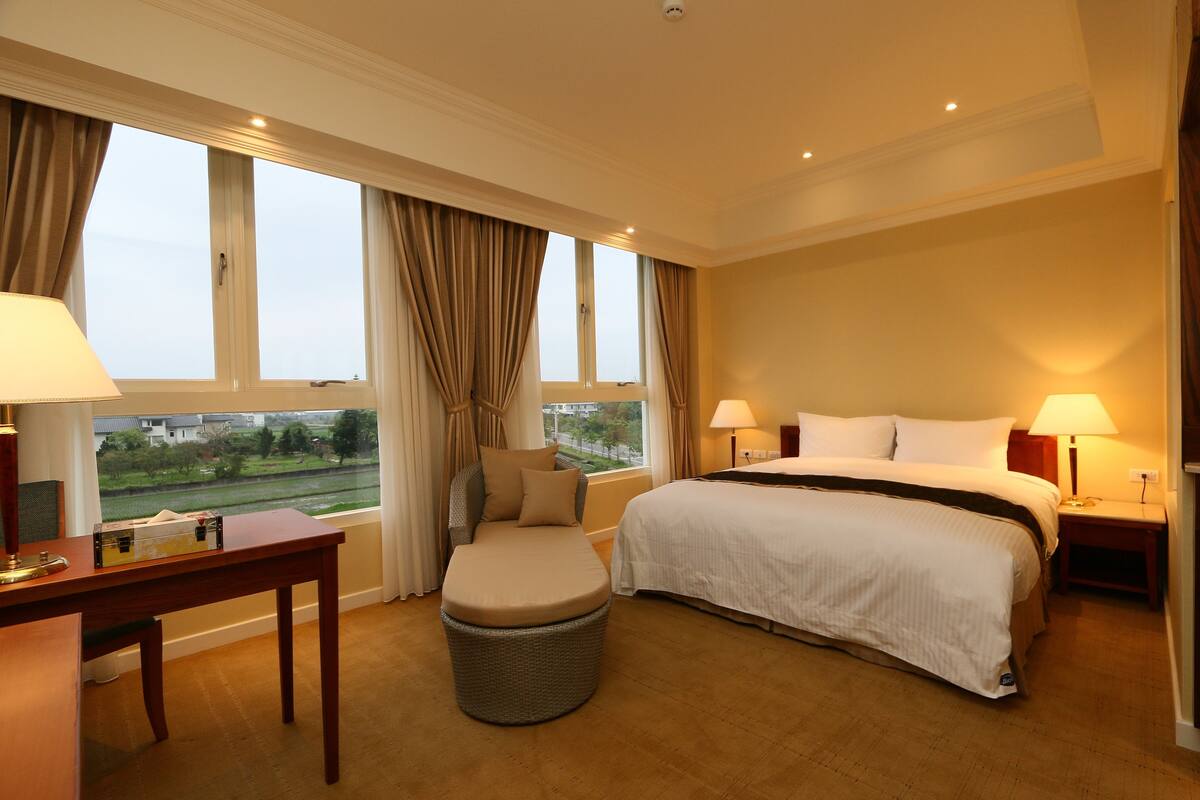 A hotel room with a large bed, a desk, a chair, and a view of the outdoors.