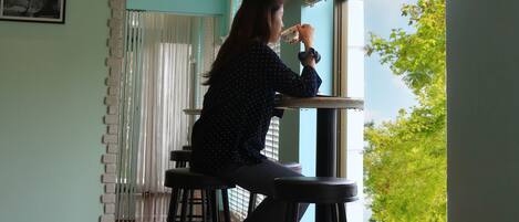 A person sitting at a table by a window with blinds, in a room with wooden flooring and a light blue wall.