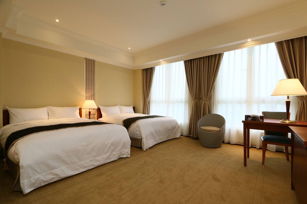 A hotel room with two beds, a desk, a chair, and a window with curtains.