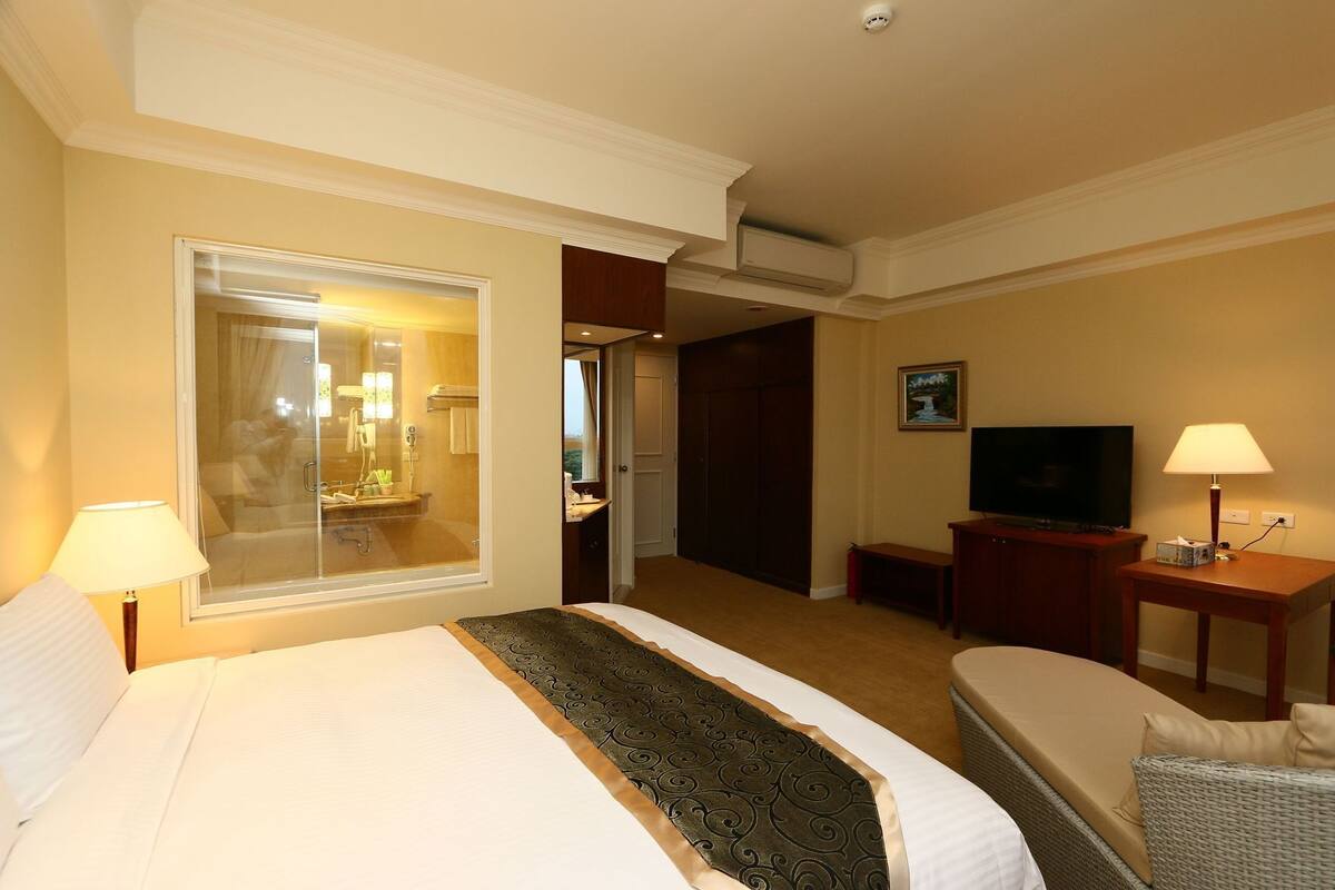 A hotel room with a bed, a television, a desk with a lamp, a chair, and a bathroom visible through a glass door.
