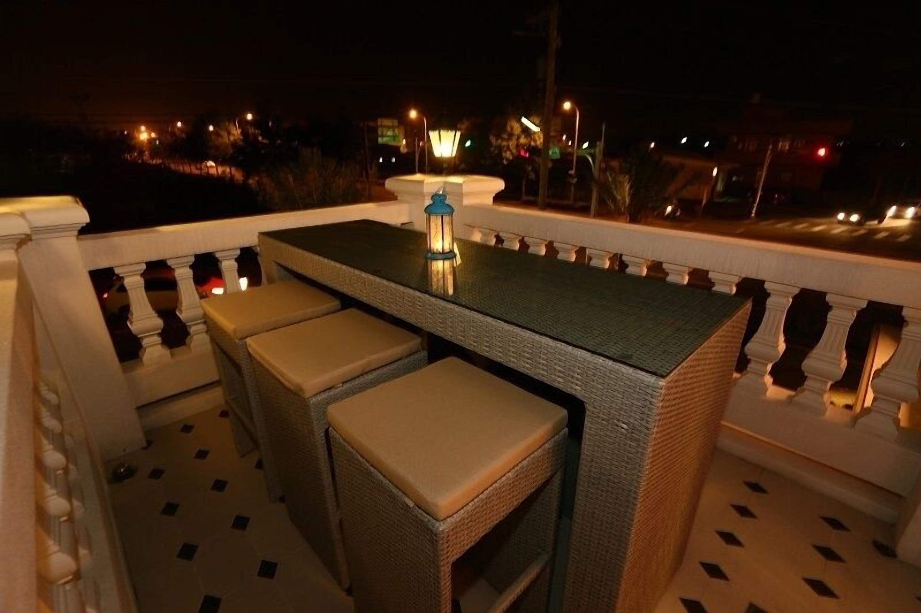 Un balcón con muebles de mimbre y una mesa, con un farol y vista a un paisaje urbano de noche.