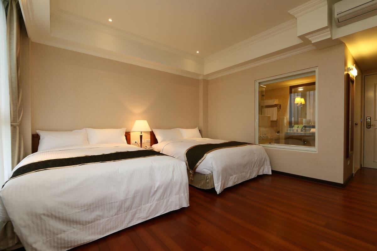 A hotel room with two beds, wooden flooring, a large mirror, and a window.
