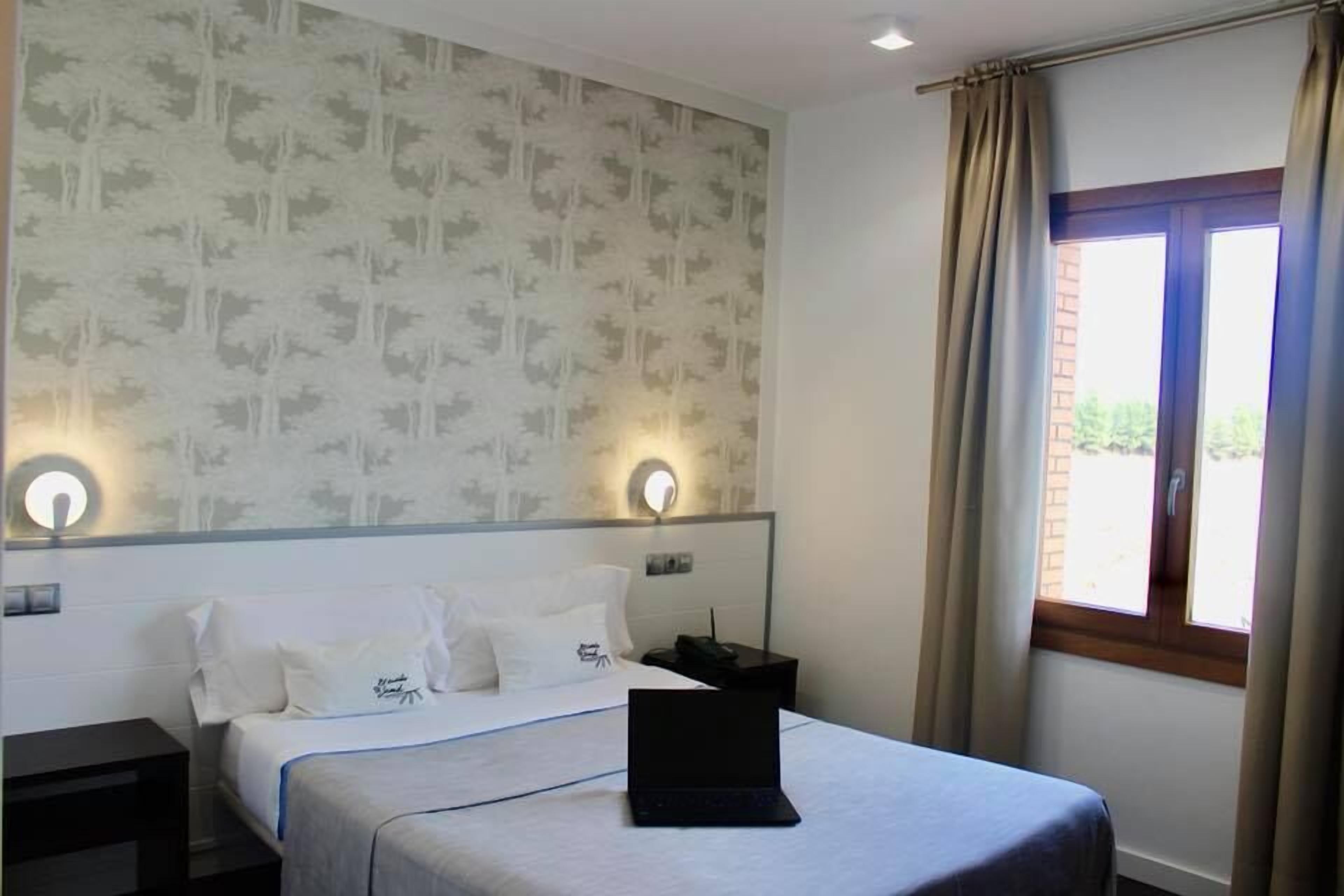 Una habitación de hotel con una cama, una computadora portátil sobre la cama, una mesita de noche, una pared con papel tapiz estampado y una ventana con cortinas.