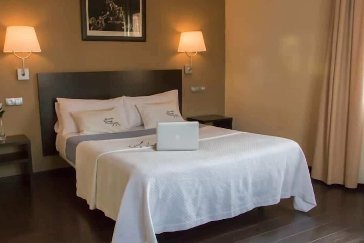 Una cama bien hecha con una computadora portátil, dos lámparas de noche y un cuadro en la pared.