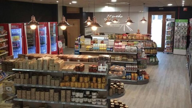 Interior de una tienda de comestibles con dispensadores refrigerados para bebidas, estantes llenos de refrigerios y luces colgantes.