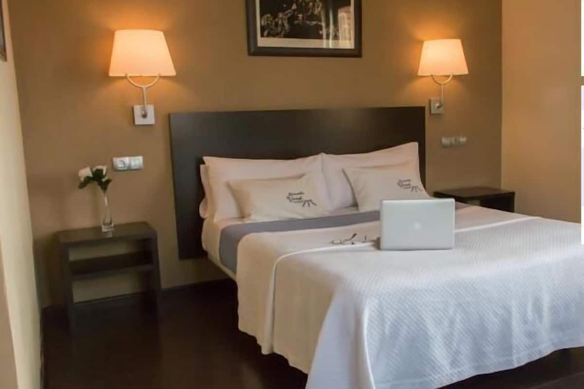 Habitación de hotel con cama, dos lámparas de noche, una computadora portátil sobre la cama, mesita de noche con un florero y un cuadro en la pared.
