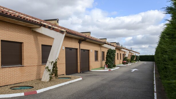 A row of brick buildings with brown doors and windows, a clear sky, and a road with directional arrows. - Motel Cancun León (Ardon)