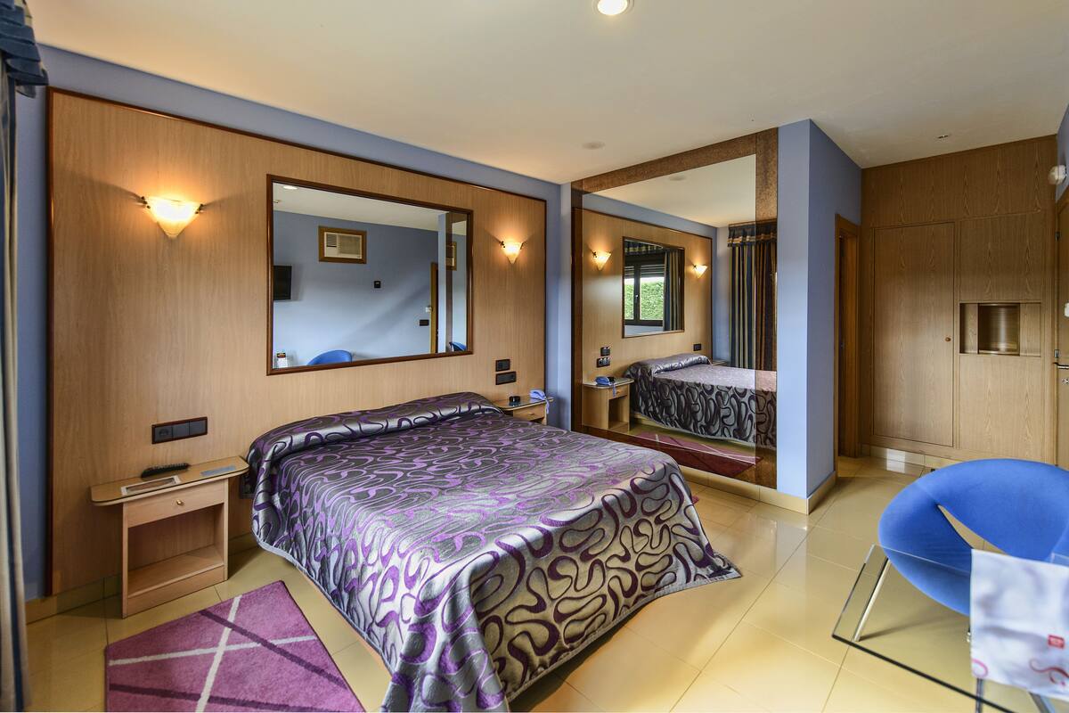 A modern hotel room with a large bed, a desk, a chair, and a mirror.