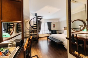 A hotel room with a wooden floor, a bed, a desk with a chair, a mirror, a window with curtains, and a spiral staircase.