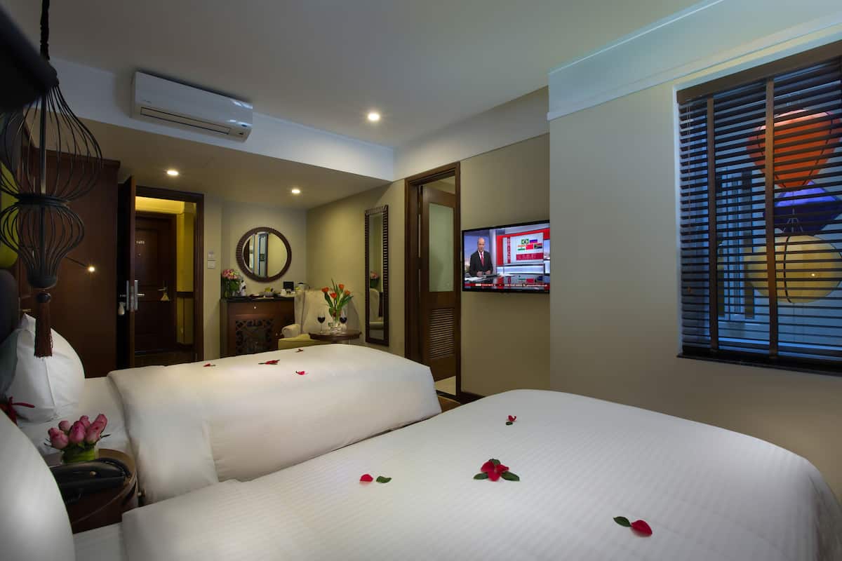 A hotel room with two beds, a television, a mirror, and a window with blinds.