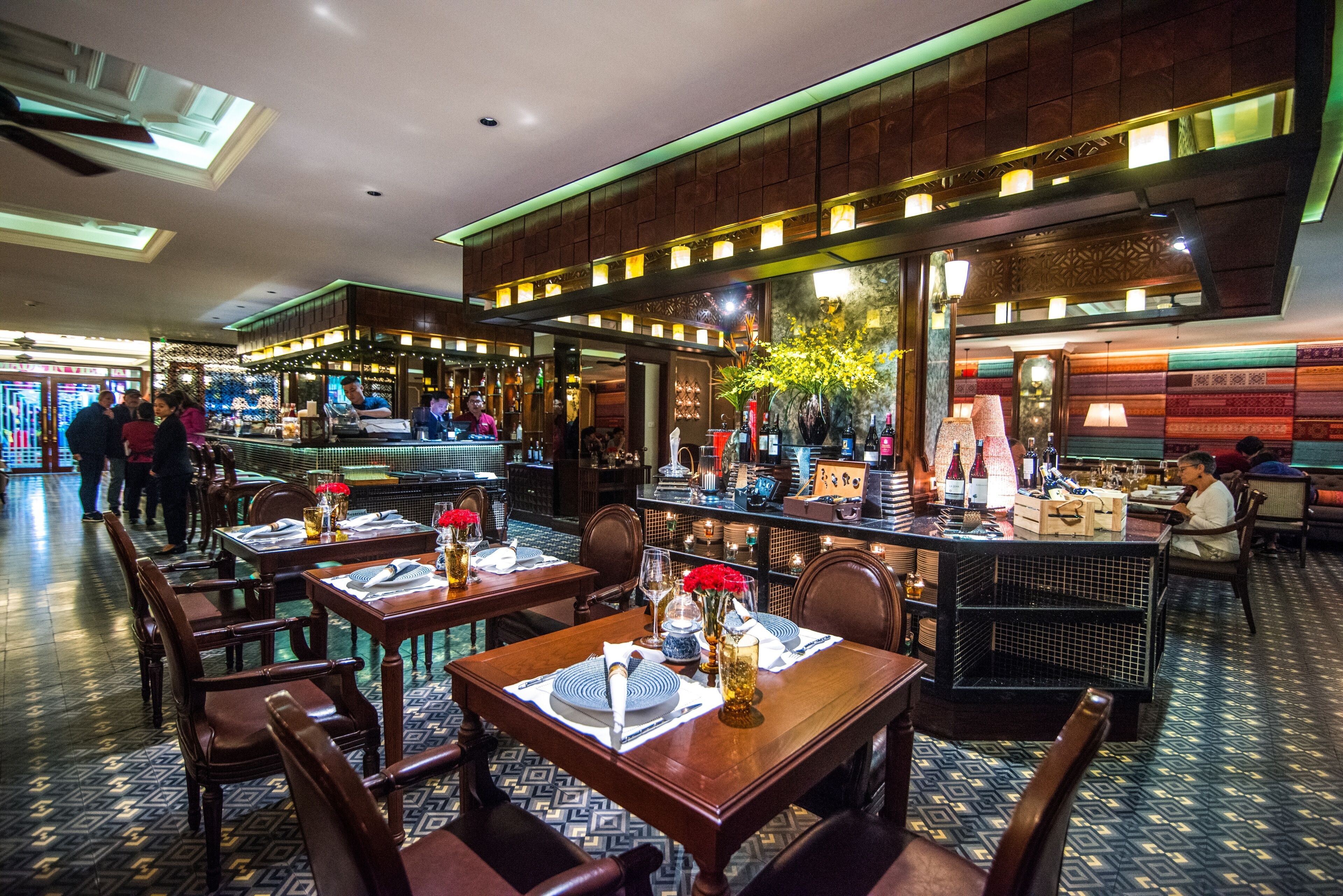 A restaurant with multiple tables set for dining, a bar area, and a visible kitchen in the background.