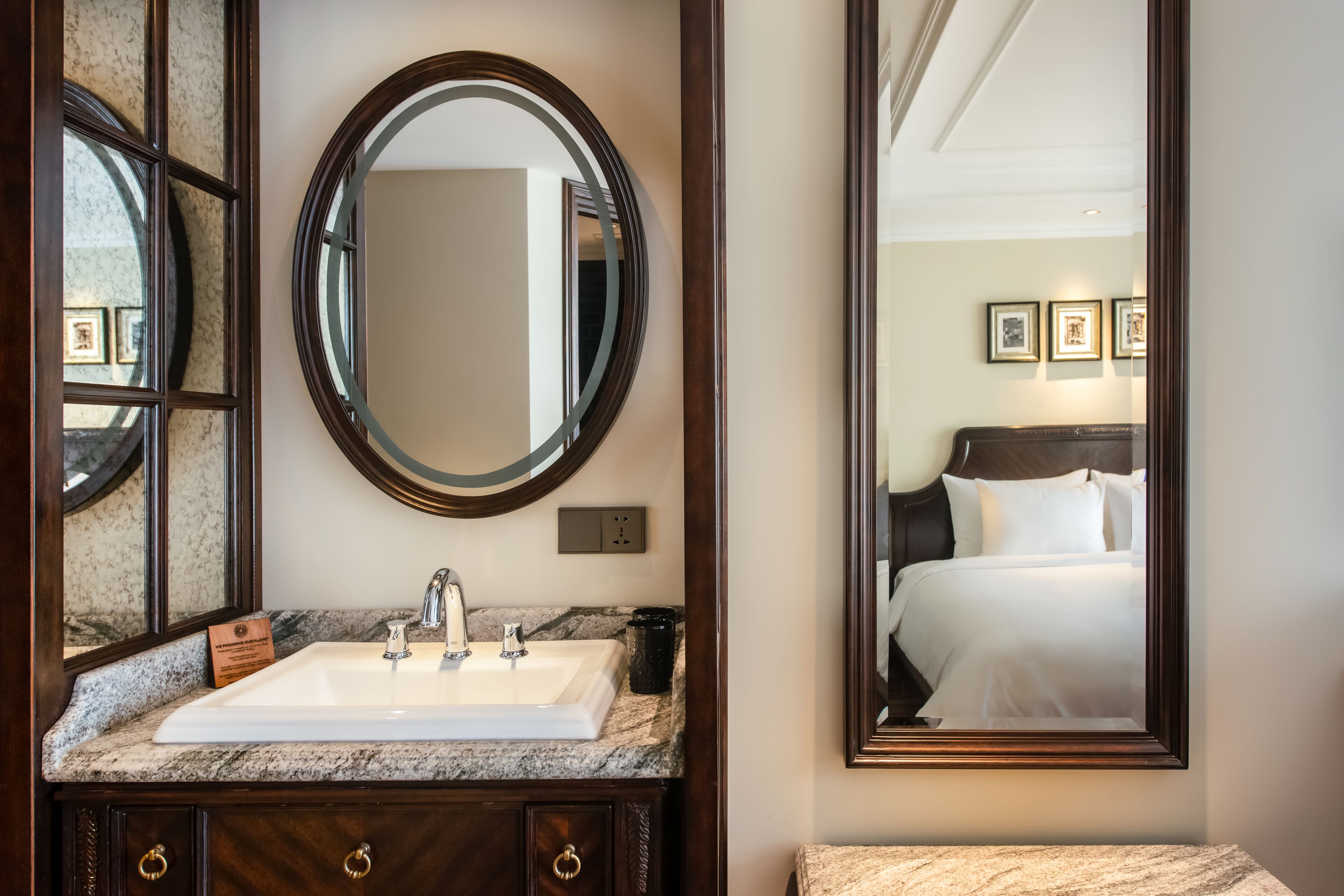 A bathroom with a marble sink, a large mirror, and a framed bed reflection.