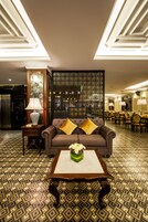 Un vestíbulo de hotel con suelo estampado, un sofá marrón con cojines amarillos, una mesa con un jarrón de flores y una pared decorativa con patrones geométricos.