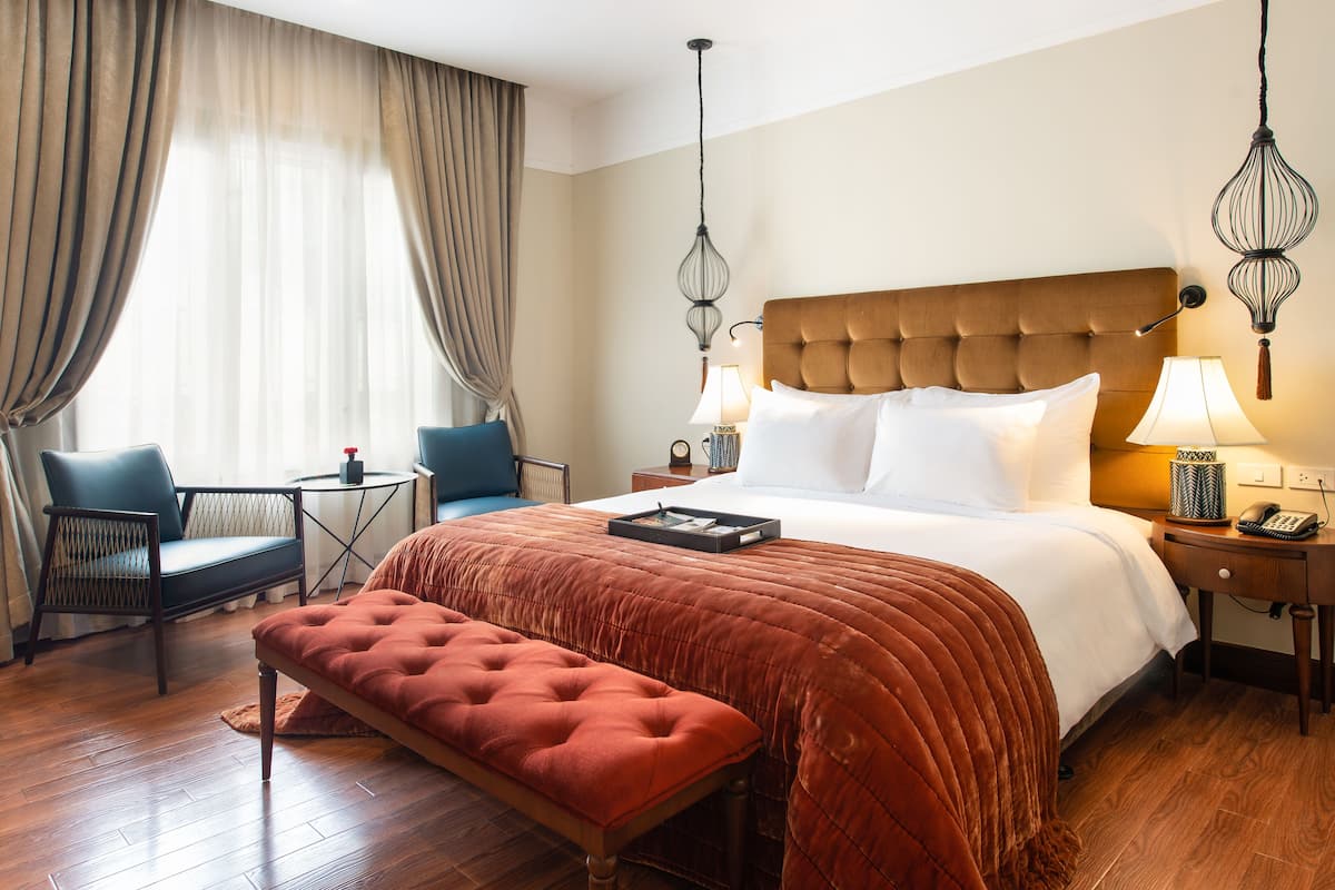 A hotel room with a large bed, a bench, a chair, a small table, and a lamp.