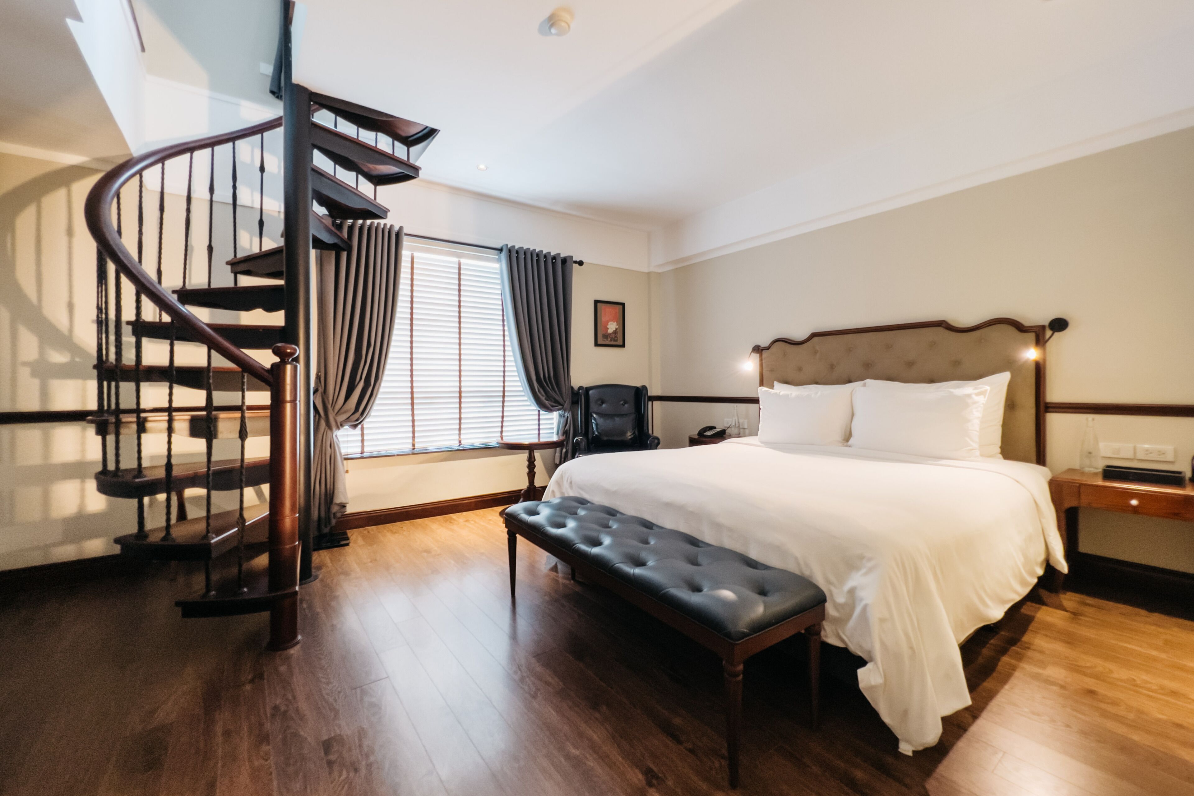 Een slaapkamer met een groot bed, een houten bankje, een bureau en een spiltrap.