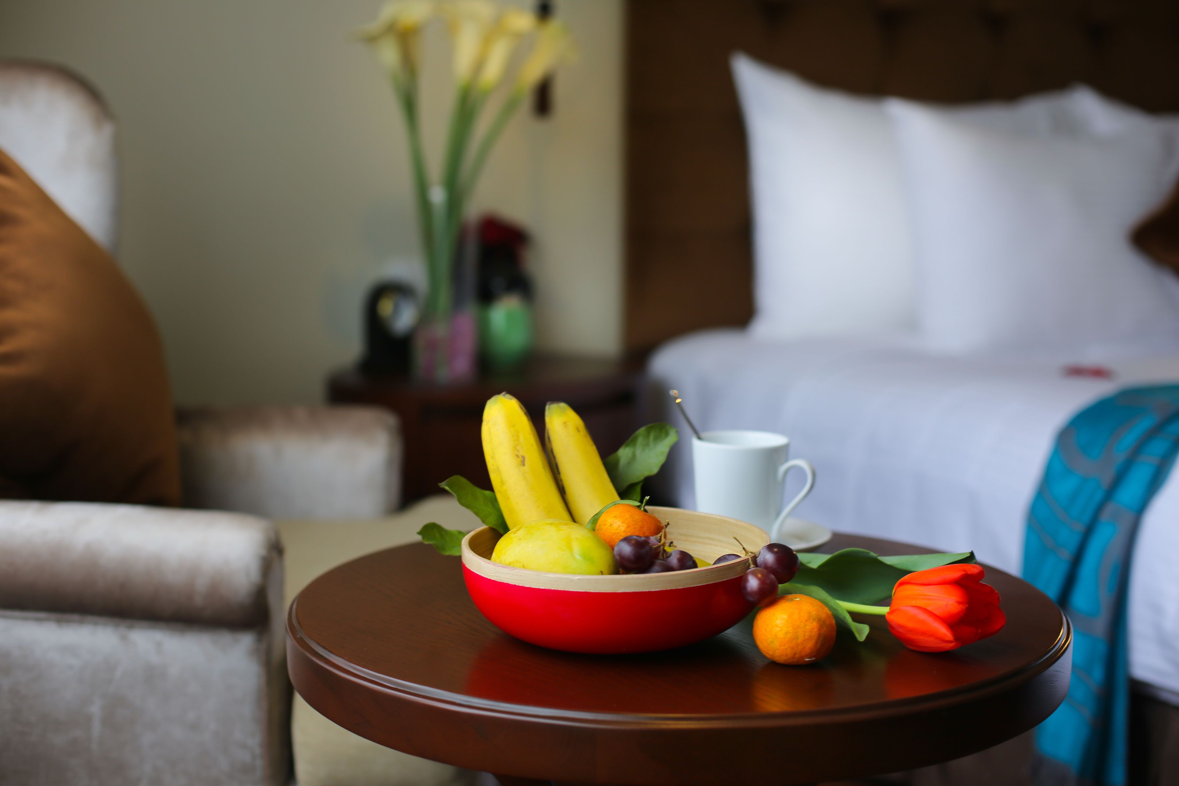 A hotel room with a bed, a bedside table with a bowl of fruit, a white mug, and a vase with flowers.