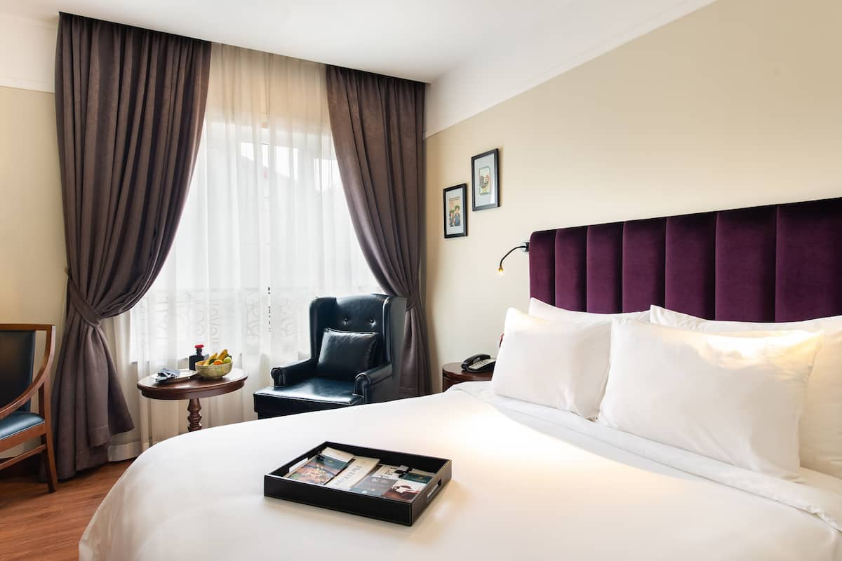 A hotel room with a large bed, a chair, a small table with a tray of items, and a window with curtains.