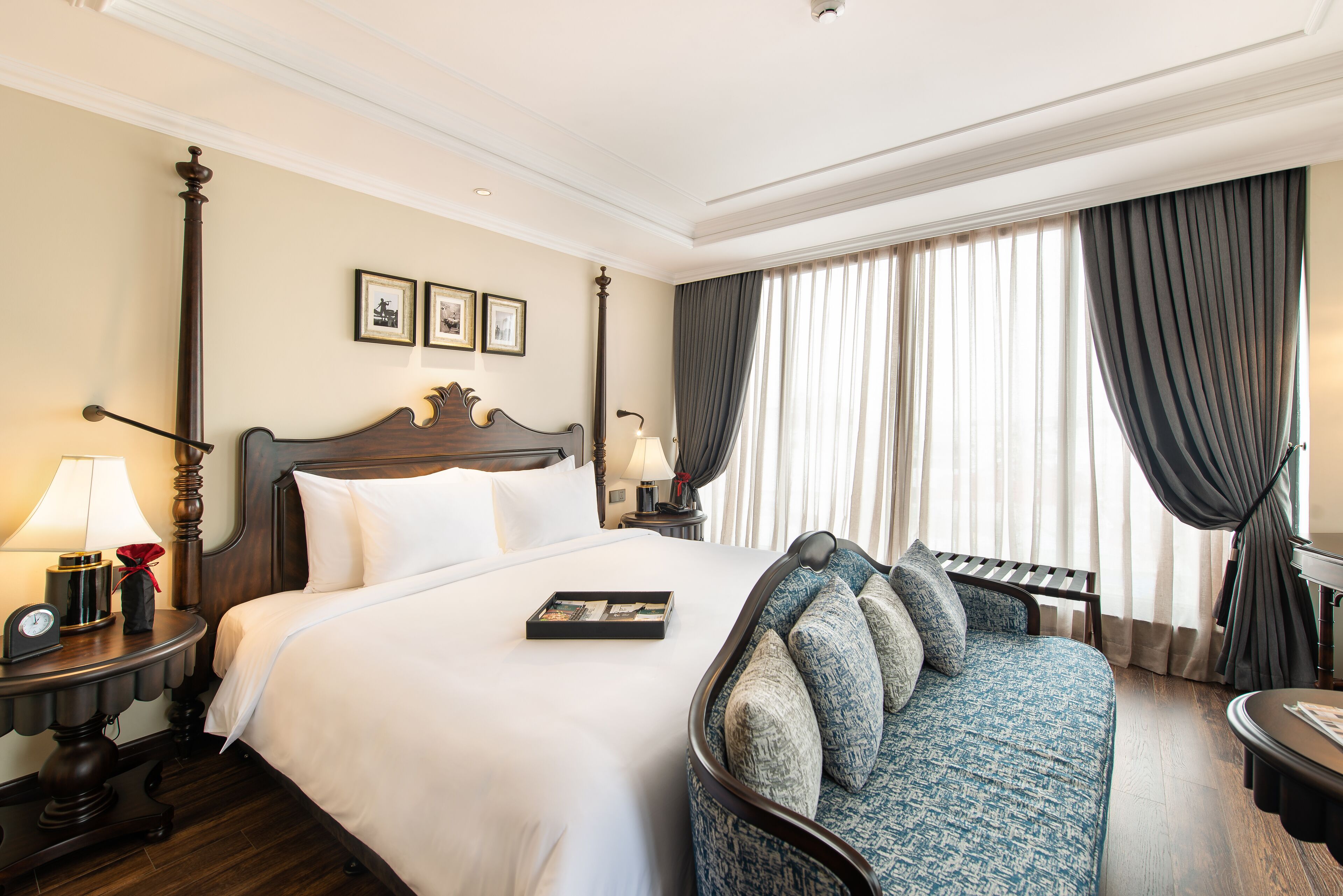 A hotel room with a large bed, a sofa, a nightstand with a lamp, and a window with curtains.