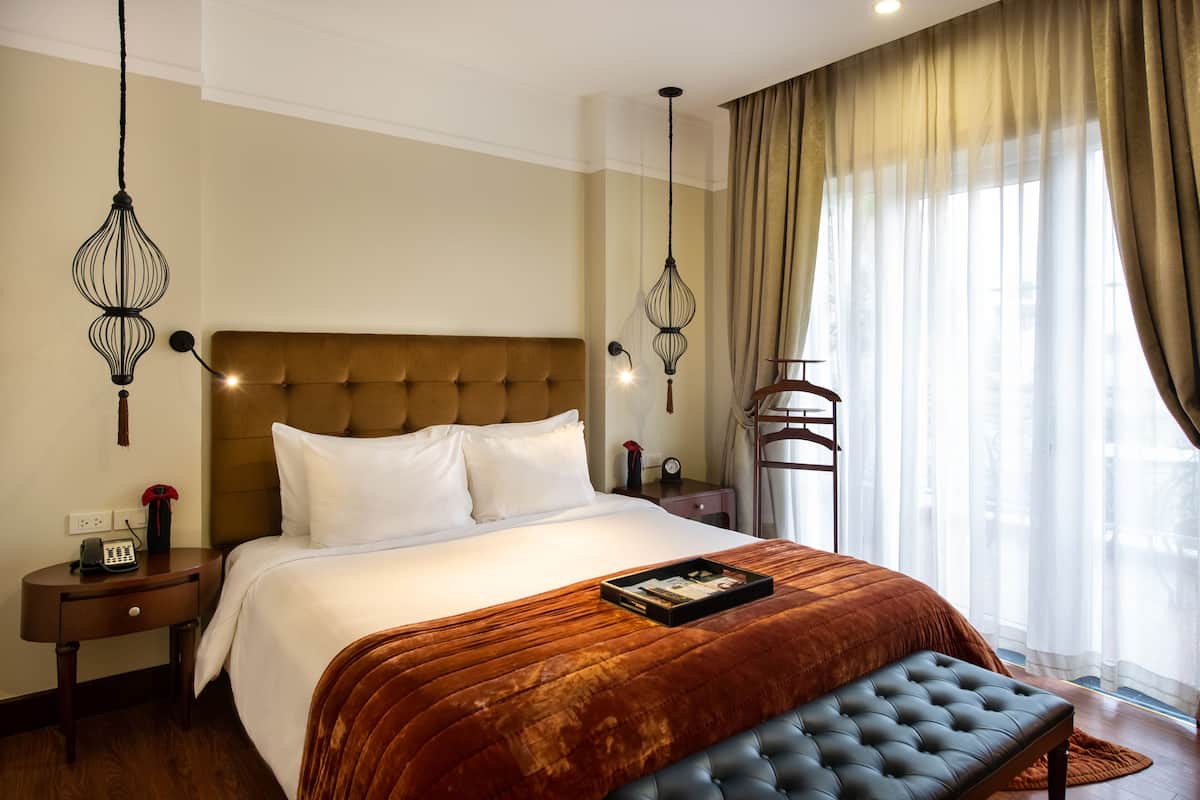 A hotel room with a large bed, a bedside table with a telephone, a bedside lamp, and a window with curtains.
