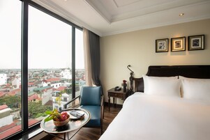 A hotel room with a large window, a bed, a chair, a small table with a fruit bowl, and framed pictures on the wall.