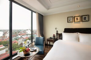 Enjoy a panoramic city view from the comfort of this stylish room, featuring a plush bed, a cozy chair, and a small table with a fruit bowl.