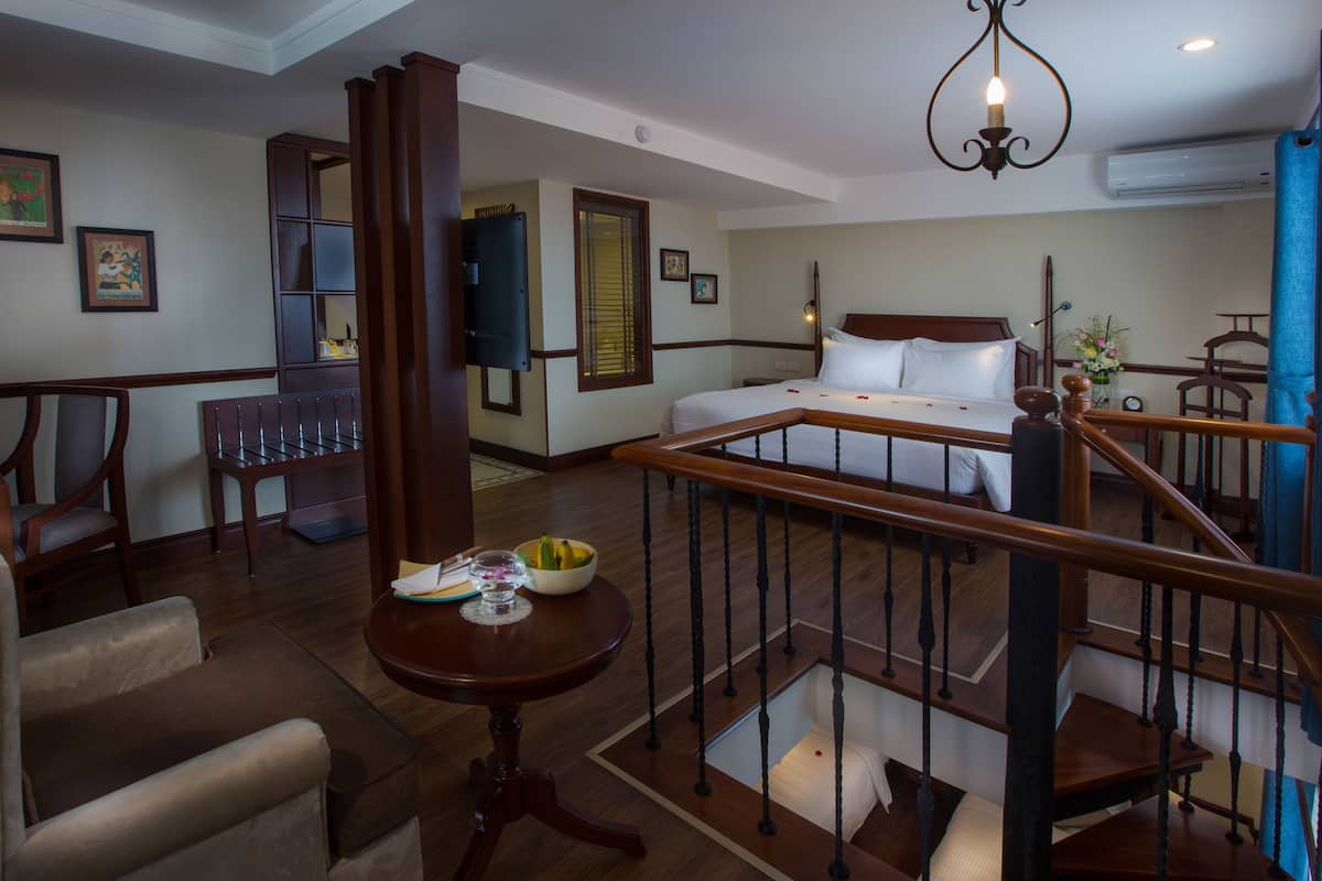 A hotel room with a bed, a desk, a chair, a table with a bowl of fruit, and a staircase.
