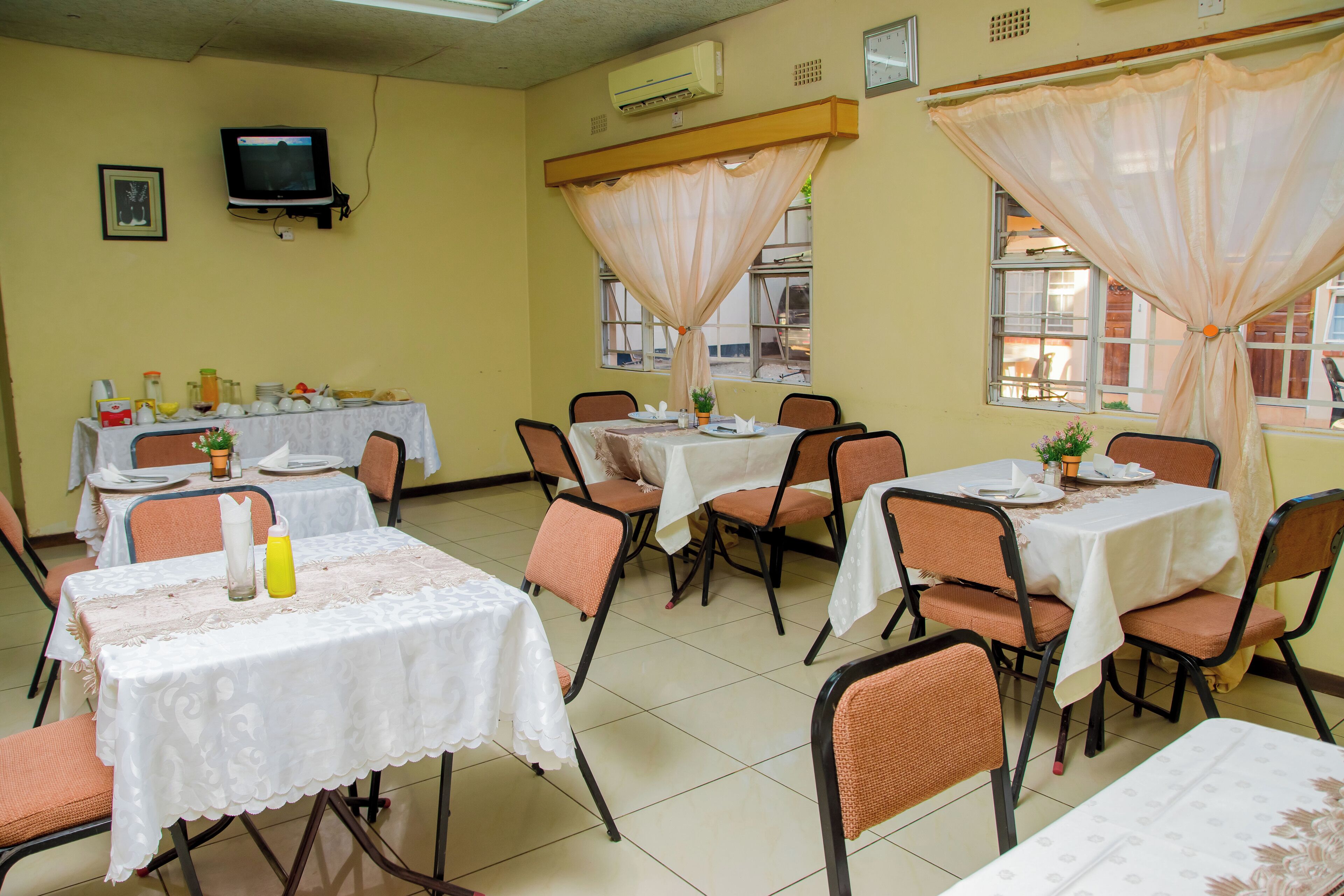 Un comedor con mesas dispuestas para comidas, sillas, un televisor en la pared y ventanas con cortinas.