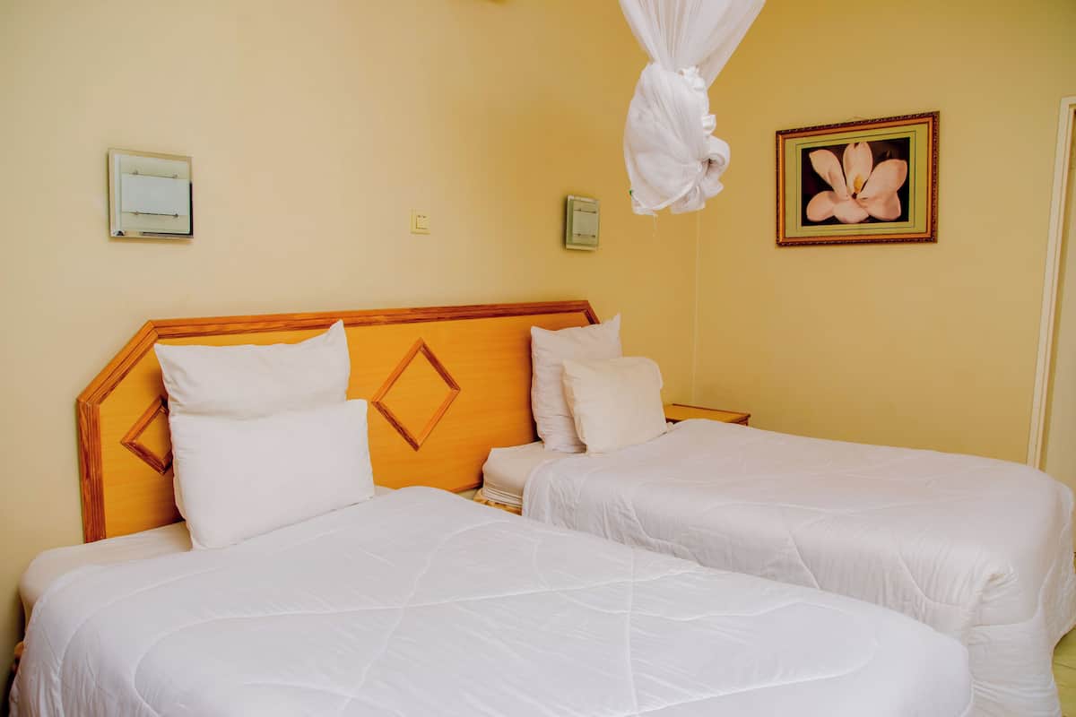 A hotel room with two beds, a wooden headboard, white bedding, a wall-mounted air purifier, a framed picture, and a hanging mosquito net.